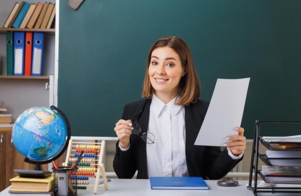 young-woman-teacher-wearing-glasses-sitting-school-desk-with-globe-books-front-blackboard-classroom-holding-white-empty-sheet-paper-happy-positive-smiling_141793-133110